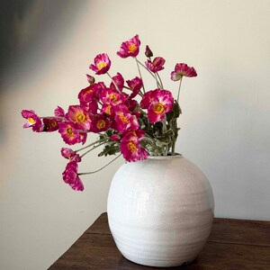 May include: A white ceramic vase with a round, textured body, filled with a bouquet of pink poppy flowers.