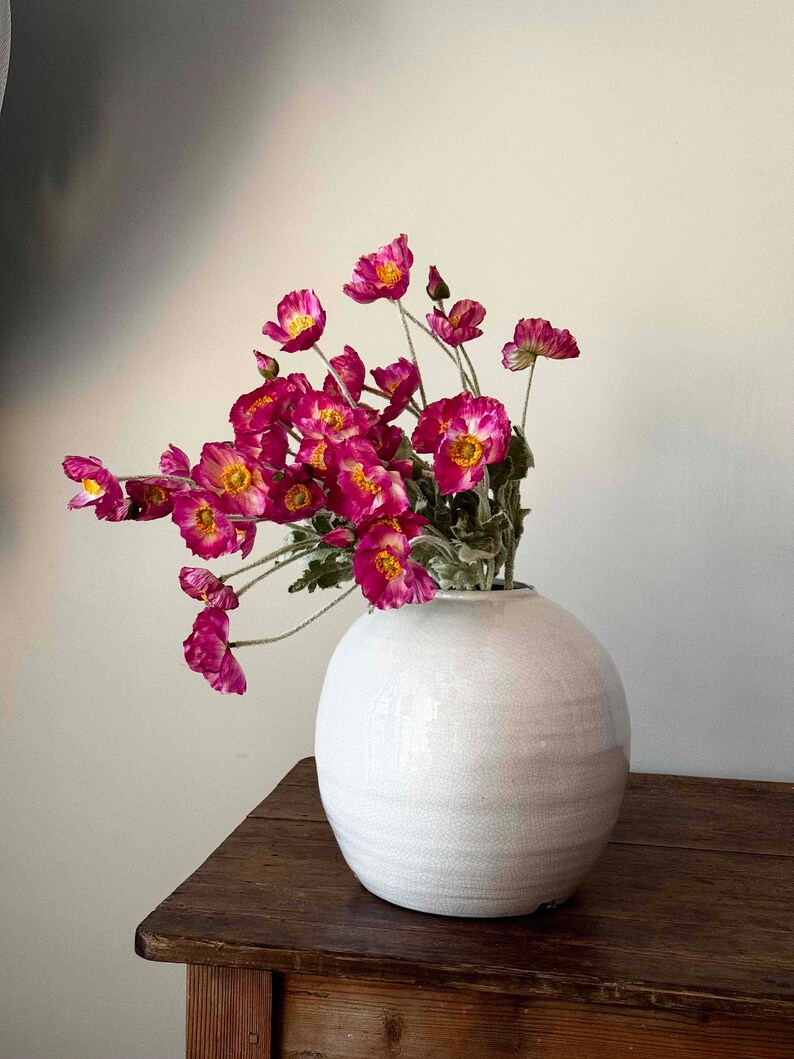 May include: A white ceramic vase with a round, textured body, filled with a bouquet of pink poppy flowers.