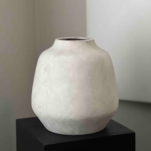 May include: A white ceramic vase with a rounded, slightly flared opening and a smooth, textured surface. The vase is sitting on a black pedestal.