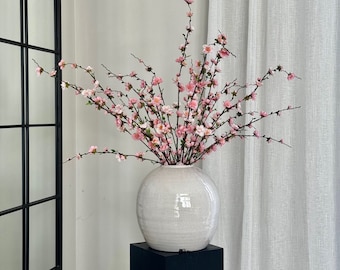 May include: A white ceramic vase with a textured surface holds a bouquet of pink cherry blossom branches. The vase is sitting on a black pedestal.