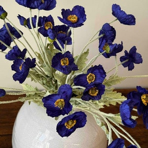 May include: A bouquet of deep blue silk poppy flowers with yellow centers arranged in a white ceramic vase with a crackled glaze.
