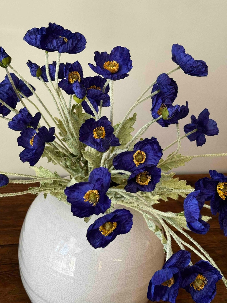 May include: A bouquet of deep blue silk poppy flowers with yellow centers arranged in a white ceramic vase with a crackled glaze.