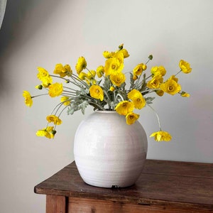 May include: A bouquet of yellow flowers in a white ceramic vase on a wooden table.