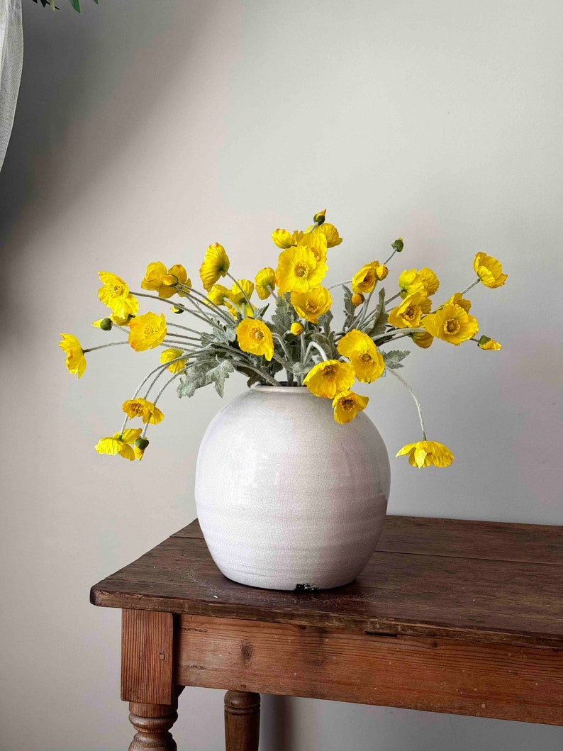 May include: A bouquet of yellow flowers in a white ceramic vase on a wooden table.