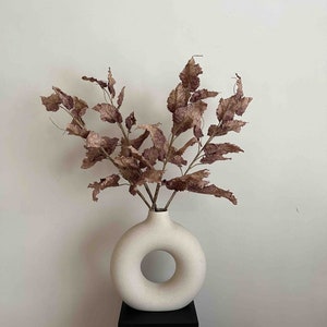 May include: A white ceramic vase with a donut-shaped opening, holding dried brown leaves. The vase is on a black pedestal.