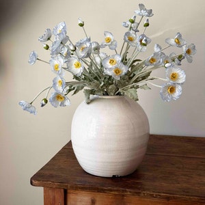 May include: A bouquet of light blue poppy flowers in a white ceramic vase with a textured surface. The vase is sitting on a wooden table.
