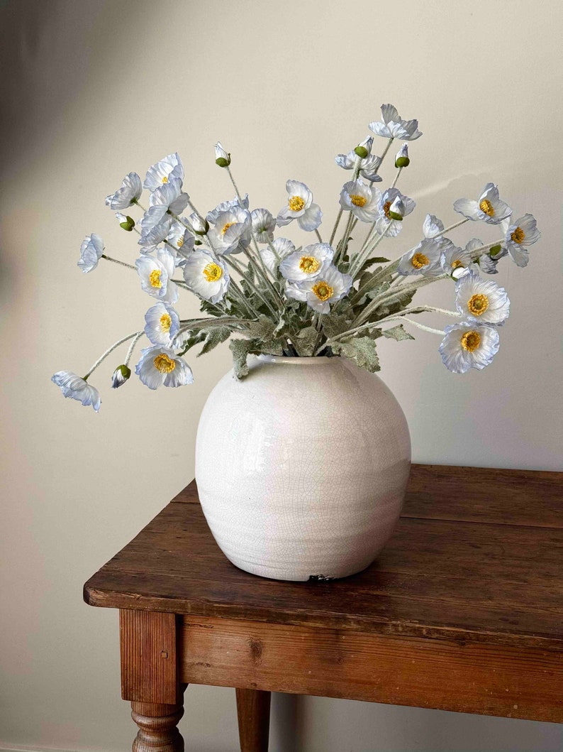 May include: A bouquet of light blue poppy flowers in a white ceramic vase with a textured surface. The vase is sitting on a wooden table.