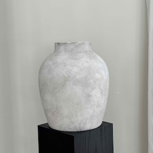 May include: A white ceramic vase with a textured surface, sitting on a black wooden plinth.