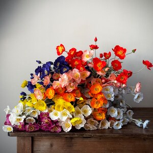 May include: A bouquet of silk poppy flowers in various colors, including red, orange, yellow, white, pink, blue, and purple. The flowers are arranged on a wooden table.
