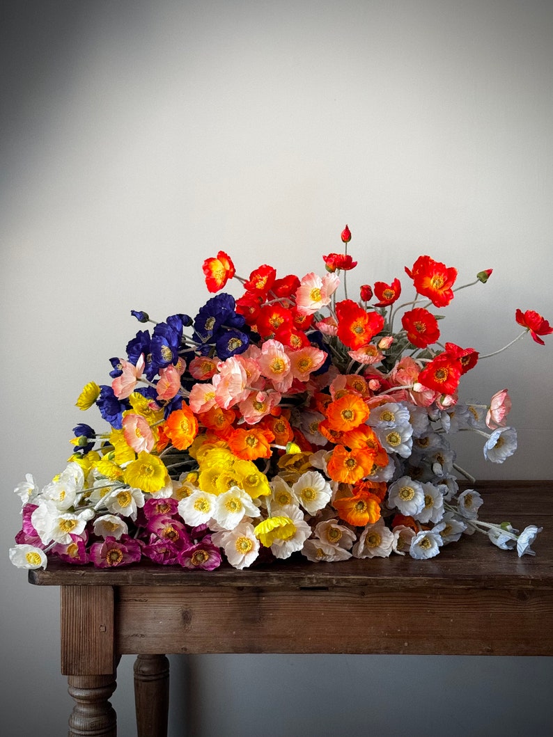 May include: A bouquet of silk poppy flowers in various colors, including red, orange, yellow, white, pink, blue, and purple. The flowers are arranged on a wooden table.