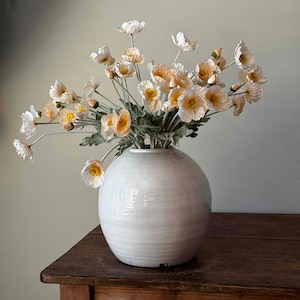 May include: A white ceramic vase with a round, slightly textured body, filled with a bouquet of delicate, pale pink and white poppy flowers with yellow centers.