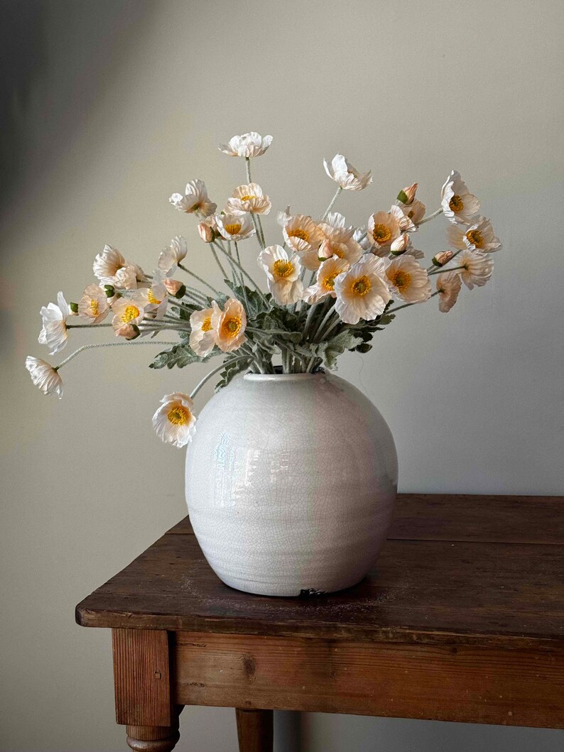 May include: A white ceramic vase with a round, slightly textured body, filled with a bouquet of delicate, pale pink and white poppy flowers with yellow centers.