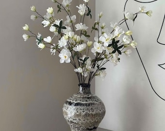May include: A decorative vase with a speckled, metallic finish holds a bouquet of white flowers with green leaves. The vase is placed on a black square pedestal. The flowers have delicate white petals and are in full bloom.