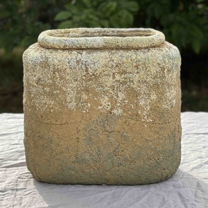 May include: A square, textured, concrete planter with a light brown and green colour scheme. The planter has a slightly rounded top edge.