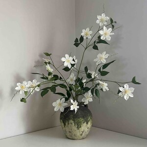 以下が含まれることがあります： 緑色の釉薬がかかった白い陶器の花瓶に、白い花と緑の葉が活けられています。花瓶は素朴で古びた風合いをしています。