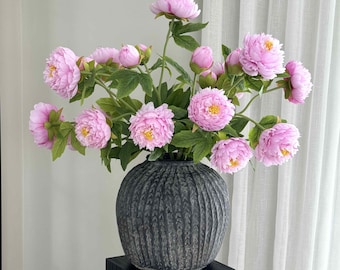 May include: A bouquet of pink peonies with yellow centers and green leaves in a dark gray, textured vase. The vase sits on a black square pedestal, with a white curtain in the background. The flowers are in full bloom.