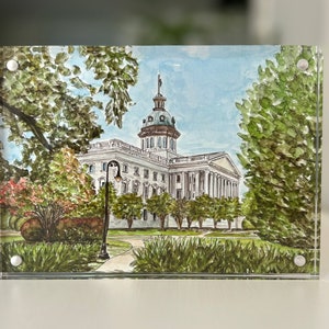 May include: Watercolor painting of a white building with a domed roof, framed by lush green trees and bushes. The artwork is displayed in a clear acrylic block, showcasing the architectural details and surrounding landscape. A path leads to the building.