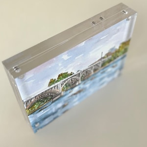 K&ouml;nnte beinhalten: Ein Aquarellgem&auml;lde einer Br&uuml;cke &uuml;ber einem Fluss, eingefasst in einem transparenten Acrylblock. Das Kunstwerk zeigt einen blauen Fluss, eine graue Br&uuml;cke mit B&ouml;gen und gr&uuml;ne B&auml;ume. Der Acrylblock ist rechteckig und hat silberne Kanten.