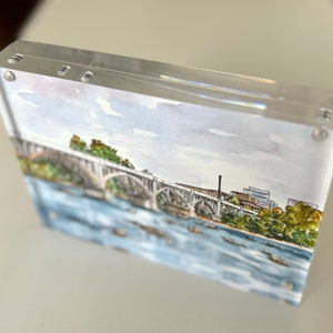 K&ouml;nnte beinhalten: Aquarellmalerei einer Br&uuml;cke &uuml;ber einen Fluss, pr&auml;sentiert in einem transparenten Acrylrahmen. Das Kunstwerk zeigt eine detaillierte Darstellung der Br&uuml;cke mit gr&uuml;nen B&auml;umen und einem blauen Himmel, der sich im Wasser spiegelt. Der Rahmen wird von kleinen Silberschrauben zusammengehalten.