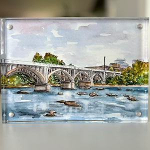 K&ouml;nnte beinhalten: Aquarellmalerei einer Br&uuml;cke &uuml;ber einem Fluss, pr&auml;sentiert in einem transparenten Acrylrahmen. Die Br&uuml;cke hat mehrere B&ouml;gen und ist in Grau- und Braunt&ouml;nen gemalt. Gr&uuml;ne B&auml;ume s&auml;umen die Ufer, der Himmel ist hellblau.