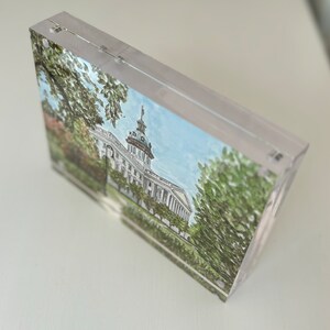 May include: A clear acrylic block featuring a watercolor painting of a white building with a dome, trees, and a blue sky. The artwork is encased in a transparent, rectangular block, showcasing the architectural illustration.