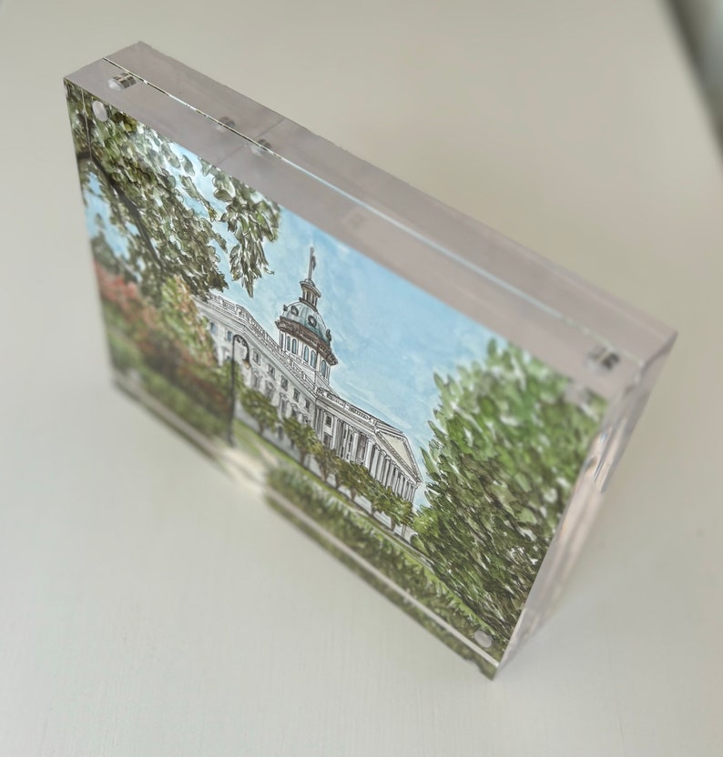 May include: A clear acrylic block featuring a watercolor painting of a white building with a dome, trees, and a blue sky. The artwork is encased in a transparent, rectangular block, showcasing the architectural illustration.