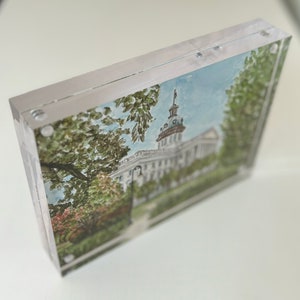 May include: A framed watercolor print of a white building with a domed roof, surrounded by trees and greenery. The artwork is encased in a clear acrylic frame, showcasing the image's details. The overall aesthetic is a blend of art and architecture.