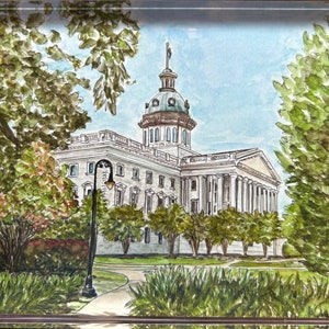 May include: Watercolor painting of a white building with a dome, framed by lush green trees and foliage. The artwork depicts a classical building with columns, a manicured lawn, and a blue sky. The image is set within a clear acrylic frame.