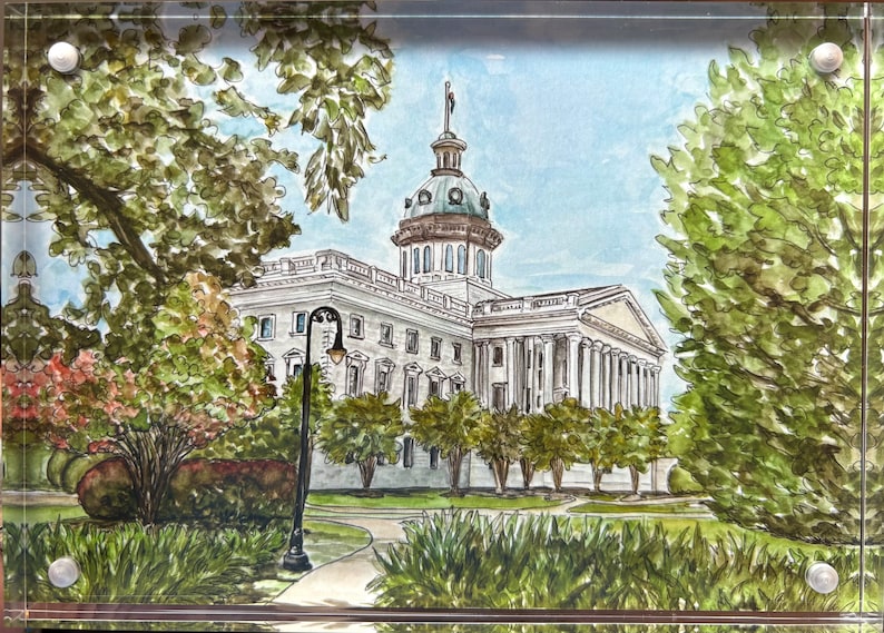 May include: Watercolor painting of a white building with a dome, framed by lush green trees and foliage. The artwork depicts a classical building with columns, a manicured lawn, and a blue sky. The image is set within a clear acrylic frame.