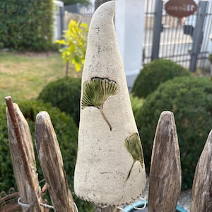 Könnte beinhalten: Eine hohe, kegelförmige Keramikskulptur mit einer strukturierten, cremeweißen Oberfläche. Das Objekt ist mit zwei geprägten, grün glasierten Ginkgoblatt-Motiven verziert. Es besitzt eine leicht gebogene, spitze Oberseite und einen flachen Boden.