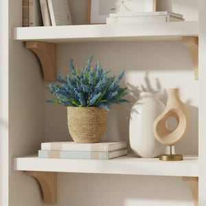 May include: A display of home decor items on white shelves. A woven basket holds blue flowers. A white vase and a wooden sculpture with a circular opening are also displayed. Books and framed art complete the arrangement.
