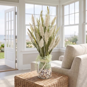 May include: A clear glass jar filled with small seashells at the base, topped with green moss and a thick rope handle. The jar contains a tall arrangement of dried cream-colored wheat stalks and slender green grass blades reaching upward.