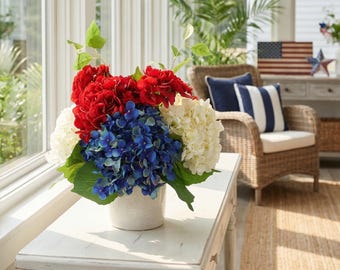 May include: A floral arrangement in a white ceramic pot features red, white, and blue flowers, including hydrangeas and other blooms. The arrangement sits on a white table in a bright room with a wicker chair and American flag decor.