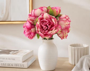 May include: A bouquet of vibrant pink and cream-colored peony flowers arranged in a simple, matte white ceramic vase. The flowers feature layered, ruffled petals with deep pink centers that transition to lighter cream edges on the outer blooms.