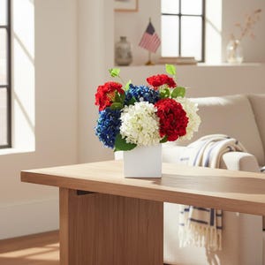 May include: A floral arrangement in a white square vase. The arrangement features red, white, and blue hydrangeas, with green leaves. The vase sits on a light brown wooden table, with an American flag in the background.