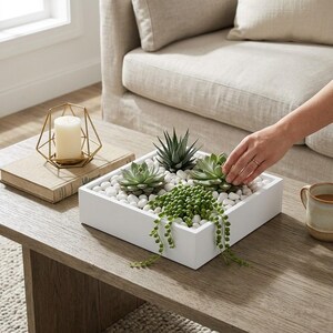 May include: A square white planter box filled with white decorative stones and three distinct succulent plants, including a trailing string of pearls, a spiky haworthia, and a rosette-shaped echeveria, arranged on a wooden surface.