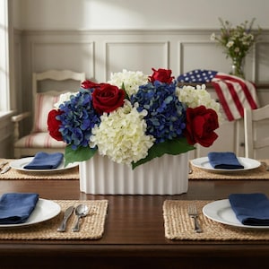May include: A floral centerpiece in a white rectangular vase, featuring red roses, blue hydrangeas, and white flowers. The arrangement sits on a dark wooden table set for a meal, with place settings and blue napkins.
