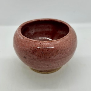 May include: A small, round, handmade ceramic bowl with a reddish-brown glaze. The bowl has a textured surface and a lighter-coloured base. The interior of the bowl is a darker shade of red. This decorative bowl is suitable for holding small items.