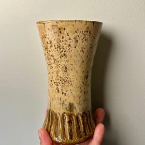May include: A handcrafted, beige ceramic vase with a textured surface and a flared top. The vase features a speckled pattern and vertical ribbed detailing at the base. The vase is held by a hand.