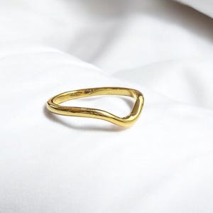 May include: A gold-colored ring with a wavy, V-shaped design. The ring is set against a white background, highlighting its smooth, reflective surface. The ring's unique shape adds a modern touch to the jewelry.