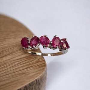 Könnte beinhalten: Ein silberner Ring mit fünf ovalen Rubin-Edelsteinen, die in einer Reihe gesetzt sind. Der Ring liegt auf einer Holzoberfläche.