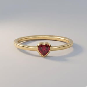 Può includere: Un anello d'oro con una gemma rosso scuro a forma di cuore. L'anello ha una fascia sottile e lucida, con la pietra a forma di cuore come elemento centrale. L'anello è posto su uno sfondo morbido e neutro.