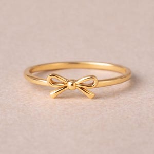 May include: A delicate gold-colored ring featuring a bow design. The ring has a thin band and a small, detailed bow at the center. The ring is set against a soft, neutral background.