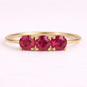 Può includere: Un anello d'oro con tre gemme rotonde di colore rosso vivo. Le gemme sono incastonate vicine, creando una linea continua di colore. La sottile fascia lucida dell'anello mette in risalto le gemme.