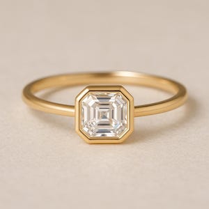 May include: A gold ring featuring a square-cut, clear gemstone. The gemstone is set within a gold bezel, and the band is a thin, polished gold. The ring is displayed on a neutral, off-white surface.