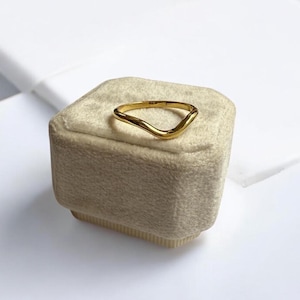 K&ouml;nnte beinhalten: Ein goldfarbener Ring mit einem einzigartigen, wellenf&ouml;rmigen Design. Der Ring besteht aus einem gl&auml;nzenden, metallischen Material und liegt auf einer beigen, samtbezogenen Schachtel. Die Form des Rings ist asymmetrisch, mit einem glatten, flie&szlig;enden Aussehen.