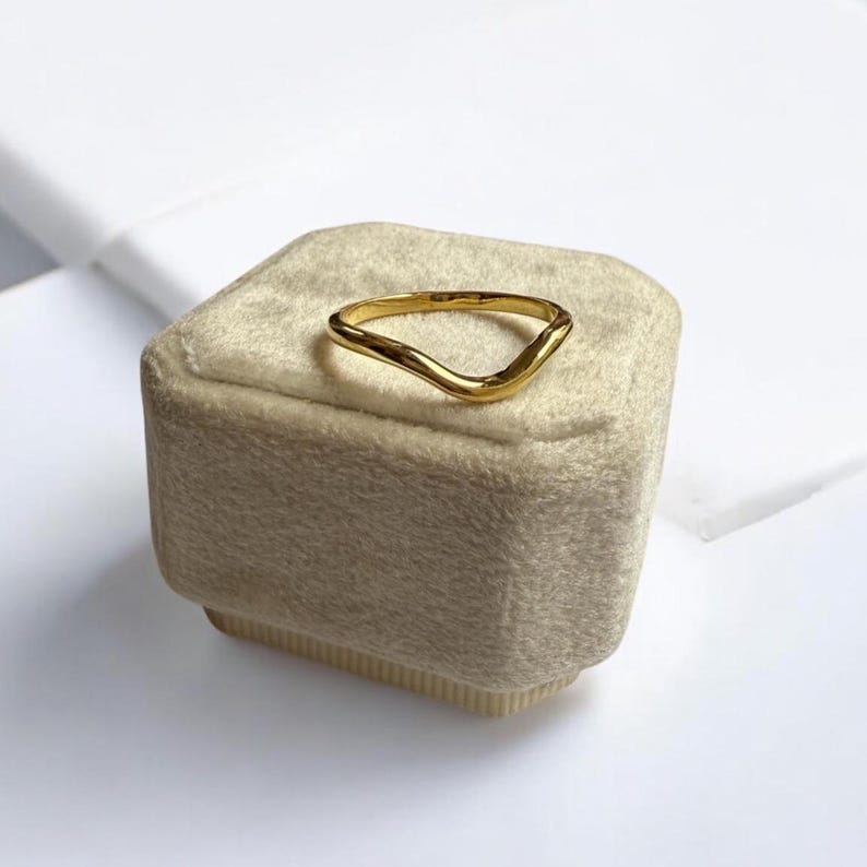 K&ouml;nnte beinhalten: Ein goldfarbener Ring mit einem einzigartigen, wellenf&ouml;rmigen Design. Der Ring besteht aus einem gl&auml;nzenden, metallischen Material und liegt auf einer beigen, samtbezogenen Schachtel. Die Form des Rings ist asymmetrisch, mit einem glatten, flie&szlig;enden Aussehen.