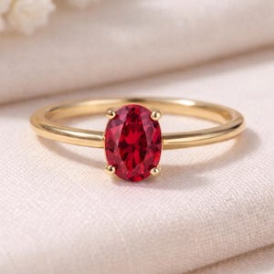 Puede incluir: Un anillo de oro con una vibrante gema roja ovalada. El anillo tiene una banda delgada y una montura de cuatro puntas que sujeta la gema. El anillo se exhibe sobre una tela suave de color blanquecino.