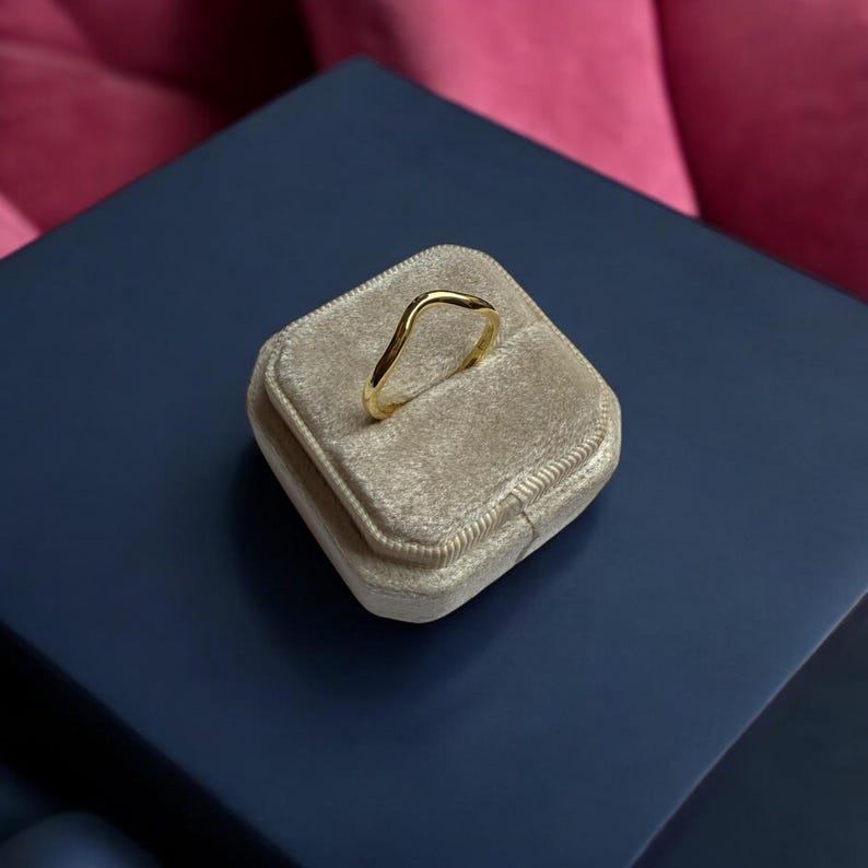 K&ouml;nnte beinhalten: Ein goldfarbener Ring mit wellenf&ouml;rmigem Design, pr&auml;sentiert auf einer beigefarbenen Samt-Ringbox. Die Ringbox befindet sich auf einer dunkelblauen Oberfl&auml;che, mit einem rosa Hintergrund. Die einzigartige Form des Rings verleiht dem Schmuck eine moderne Note.
