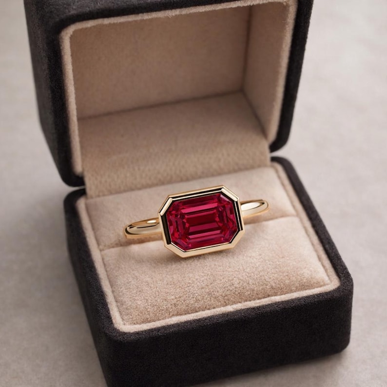 Pode incluir: Um anel de ouro com uma grande pedra preciosa retangular vermelho rubi. O anel &eacute; cravejado em uma moldura de ouro e &eacute; exibido em uma caixa de anel de veludo preto com interior bege. O design do anel &eacute; simples, mas elegante.